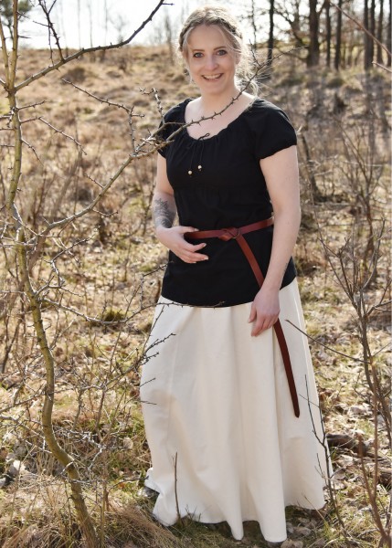 Eine Frau trägt einen mittelalterlichen, weit ausgestellten Rock in Naturfarbe. Sie steht in einem waldähnlichen Gebiet, gekleidet in einem schwarzen Oberteil und einem braunen Gürtel, der den Rock akzentuiert. Die Szene wirkt rustikal und historisch