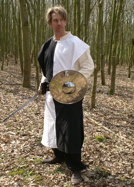 Ein Mann steht im Wald und trägt einen schwarz-weißen Wappenrock im Schachbrettmuster. Er hält ein Schwert in der rechten Hand und einen Metallschild in der linken. Der Wappenrock besteht aus robustem Stoff und bietet historische Eleganz.
